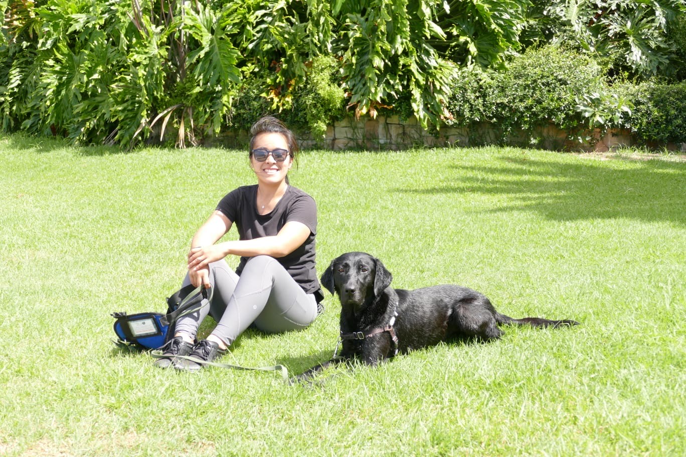 Diona sitting on the grass in the sun with Murphy, her black Labrador assistance dog, settled beside her